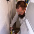 An installer wearing safety glasses and holding a four inch metal duct elbow measures the distance from a dryer outlet during a dryer vent installation in the basement of a residential home.