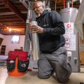 An installer kneeling on a basement floor near a furnace and water heater snaps together a two foot piece of metal piping.