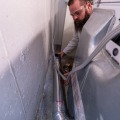 An installer wearing safety glasses and holding a four inch metal duct adjusts a section of dryer vent piping   behind an electric clothes dryer.