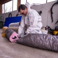 An installer wearing a white protective coverall cuts a section of of pink insulated ducting while kneeling on a concrete warehouse floor.