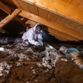 A weatherization installer kneeling in an attic with gray cellulose insulation removes a bathroom fan from between a ceiling joist cavity. 