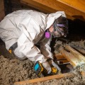A weatherization installer wearing a white protective coverall and respirator, kneels on an attic ceiling joist while preparing a drywall opening which will accommodate a new exhaust fan.