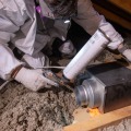 An installer wearing personal protective equipment uses a one-part foam gun to air seal a gap around a metal exhaust fan from the attic.