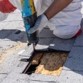 An installer kneels on an asphalt shingled roof while cutting a small hole in the roof with a reciprocal saw.