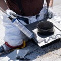 An installer kneels on an asphalt shingled roof while using a caulk gun to dispense sealant onto a black roof vent.