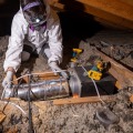 A weatherization installer wearing a head lamp and personal protective equipment kneels on an attic joist to secure a short section of metal piping to an exhaust fan.