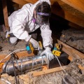 A weatherization installer wearing a head lamp and personal protective equipment kneels on an attic joist to secure a short section of metal piping to an exhaust fan with a screw gun.