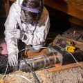 A weatherization installer wearing a head lamp and personal protective equipment kneels on an attic joist and secures a flexible duct to a section of metal ducting using a plastic zip tie. 