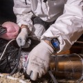 A close up image of a weatherization installer wearing personal protective equipment kneels on an attic joist and seals a joint between two pieces of ducting using a metallic foil tape. 