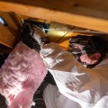 A weatherization installer wearing personal protective equipment kneels on attic joists while connecting  flexible ducting to a roof vent using a plastic zip-tie from inside an attic. 