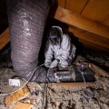 A weatherization installer wearing personal protective equipment kneels on attic joists while connecting flexible ducting using a plastic zip-tie from inside an attic. 