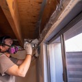 A weatherization worker applies a gray mastic sealant with a paint brush to seal the sill plate of a basement below the joist cavity.