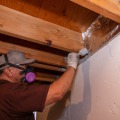 A weatherization installer wearing a baseball cap and respirator applies a gray mastic sealant to the sill plate of a home from the basement.