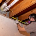 A weatherization installer wearing a respirator uses a tape measure between basement floor joists.