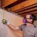 A weatherization installer wearing a respirator uses a tape measure between the subfloor and a basement sill plate.