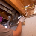 A weatherization installer wearing a respirator a piece of foil faced insulation below two white vent pipes. 