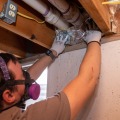 A weatherization installer wearing a respirator stuffs a piece of foil faced insulation below two white vent pipes. 