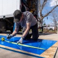 A weatherization installer kneels on a large piece of plexiglass placed on a concrete driveway to mark a measurement. 
