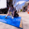 A weatherization installer wearing safety goggles holds a utility knife while kneeling on a large piece of plexiglass placed on a concrete driveway.
