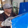 A weatherization installer wearing gloves holds a plexiglass storm window with blue protective plastic covering the window. 