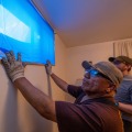 A weatherization installer holds a storm window with blue protective plastic on the glass over a window opening while inside a residential home.