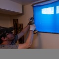 A weatherization installer uses a screw gun to install fasteners to hold a storm window with blue protective plastic on the glass over a window opening while inside a residential home.