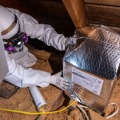 A worker wearing a white protective coverall and respirator places a pre-fabricated cover over an attic light fixture. 