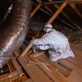A worker in an attic with a white protective coverall staples a measuring marker to attic framing.