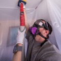 A weatherization installer within a containment work area is wearing safety goggles and respirator while holding an insulation fill tube to the ceiling.