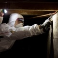 Two weatherization workers wearing protective coveralls, a respirator, and a head lamp look for air leaks in a dark crawlspace of a residential home.