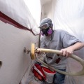 A weatherization worker wearing a respirator and safety goggles uses an tube to blow insulation into each access hole from inside a dust control enclosure.