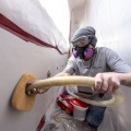 A weatherization worker wearing a respirator and safety goggles uses an tube to blow insulation into each access hole from inside a dust control enclosure.