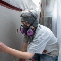 A weatherization worker wearing a respirator and safety goggles places a round two inch wooden plug into an exterior wall insulation access hole from inside a dust control enclosure.