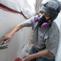 A weatherization worker wearing a respirator and safety goggles uses a hammer to tap in a round two inch wooden plug into an exterior wall insulation access hole from inside a dust control enclosure.