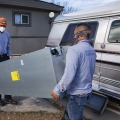 Two workers carrying a furnace into a manufactured home, both wearing a face covering.