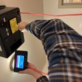 A worker using a black exhaust fan flow meter on a white ceiling exhaust panel and reading the measurements on a digital screen. 