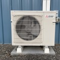 A white mini-split unit with a gray fan installed on a gray concrete pad outside a blue building. 