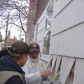 Two workers show how the white exterior cladding has been cut to allow for drill holes to be made that fit a clear fill tube to blow in gray insulation material. 