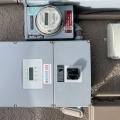 A gray photovoltaic inverter and disconnect installed on a tan textured exterior wall. 