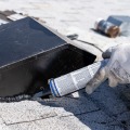 An installer seals an asphalt shingle to a black roof vent using a caulk gun to dispense sealant.