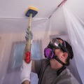 A weatherization installer within a containment work area is wearing safety goggles and respirator while holding an insulation fill tube.