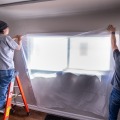 Two workers (one standing on a short ladder) hold a piece of plastic sheeting over a window from the living room of a residential home.