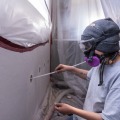 A weatherization worker wearing a respirator and safety goggles uses a plastic tie to probe a two inch round insulation access hole from inside a dust control enclosure.
