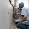 A weatherization worker wearing a respirator and safety goggles uses a putty knife to apply spackle over round two inch holes from inside a dust control enclosure.