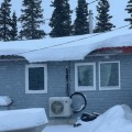 The outdoor unit of a heat pump is attached to the exterior wall of a single story home with snow on the roof.